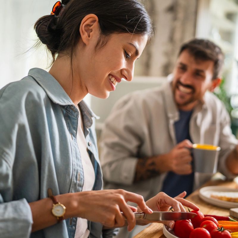 Happy young couple preparing and enjoying a healthy breakfast together, slicing fresh organic vegetables and interacting while sitting at a rustic wooden dining table in their bright kitchen