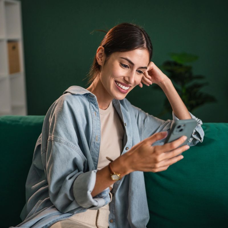 Young woman smiling and sitting comfortably on a green couch, scrolling and interacting with a smart phone, enjoying social media or online content at home