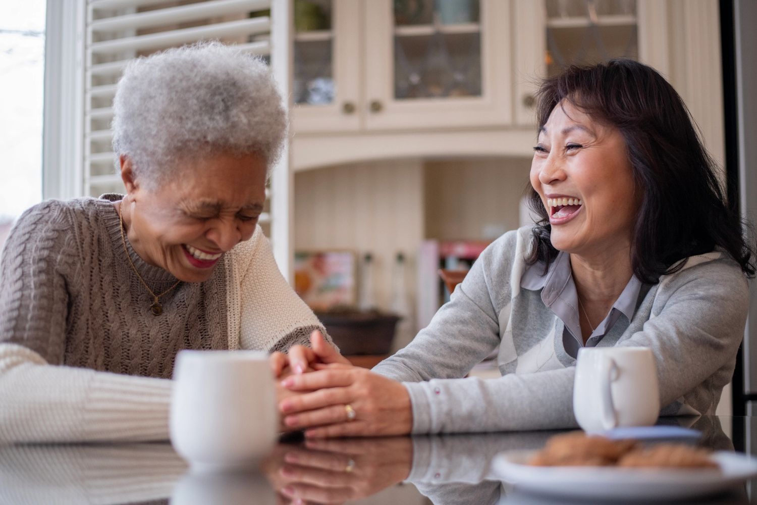 Two women laughing and holding hands over coffee at a kitchen table.