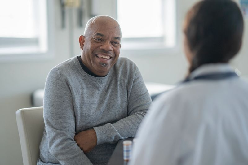 A cheerful, senior African American man sits in a clinic, sharing a warm smile with a doctor. They engage in a friendly, supportive discussion during a routine medical visit.