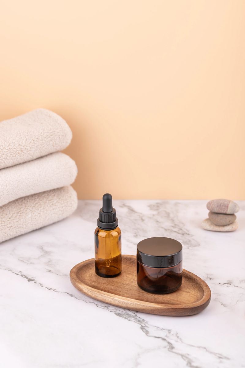 Vertical composition with amber bottle and cosmetic jar on wooden tray, beige towels and zen stones on marble. Minimal peach background. Copy space. Concept of clean beauty and wellness