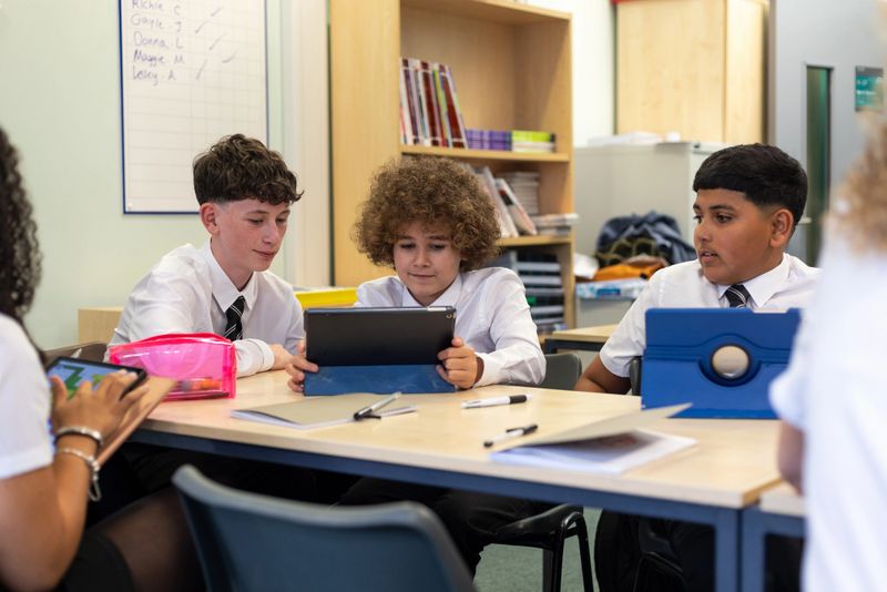 Medium shot of a group of students sitting around a table in the classroom of a school in Newcastle upon Tyne, North East England. They are focusing on digital tablets, various stationary on the table. They are wearing school uniforms.Videos similar to this scenario available.