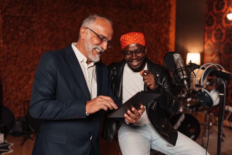 A candid interaction between two musicians, one jokingly pointing at a tablet while the other laughs, capturing the essence of camaraderie in a vibrant recording studio space.