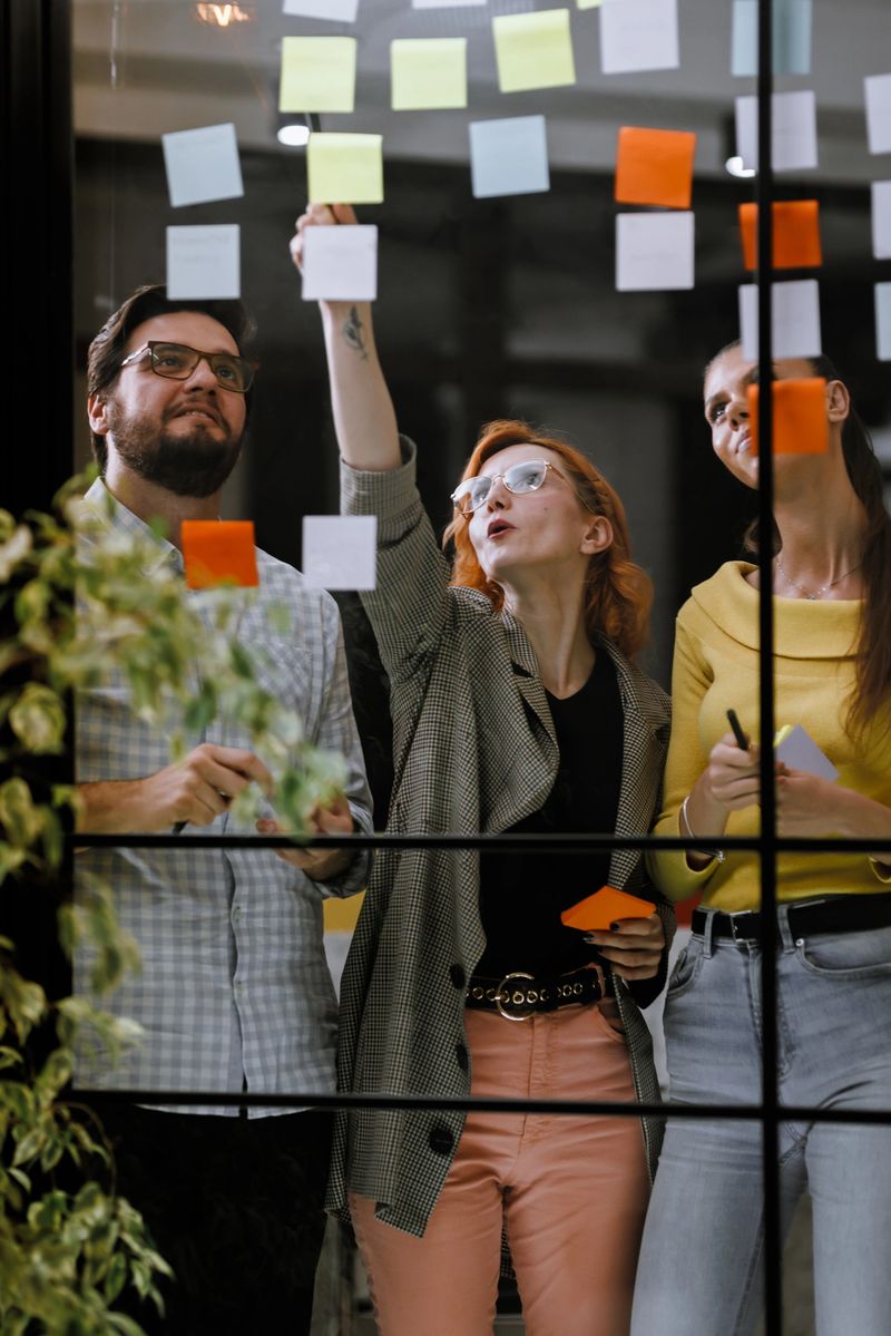 Three colleagues are engaged in a lively brainstorming session using colorful sticky notes on a glass wall in a contemporary office. They seem focused and excited about their ideas.