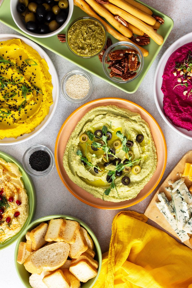 A vibrant charcuterie board features broccoli, turmeric, classic, and beetroot hummus, paired with olives, crackers, and fresh garnishes, perfect for sharing with friends