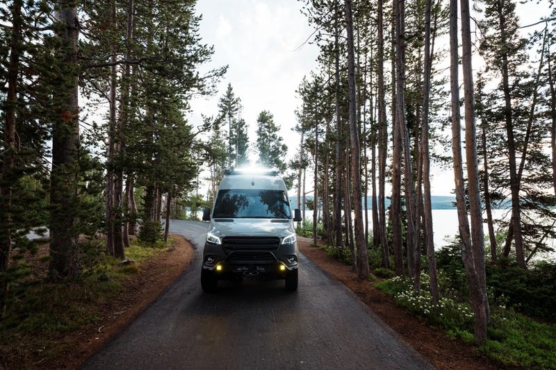 A custom build camper van is parked among towering pine trees along the shore of Lewis Lake in Yellowstone National Park, Wyoming, USA.