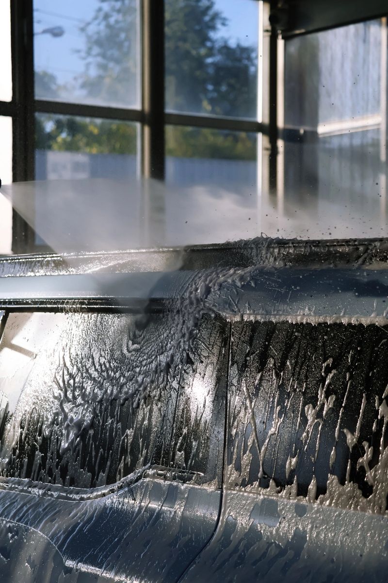 Vertical photo showing the upper part of a blue car under strong water pressure. The stream rinses white foam from the roof rail mounts, with backlight from large windows creating a bright clean mood.