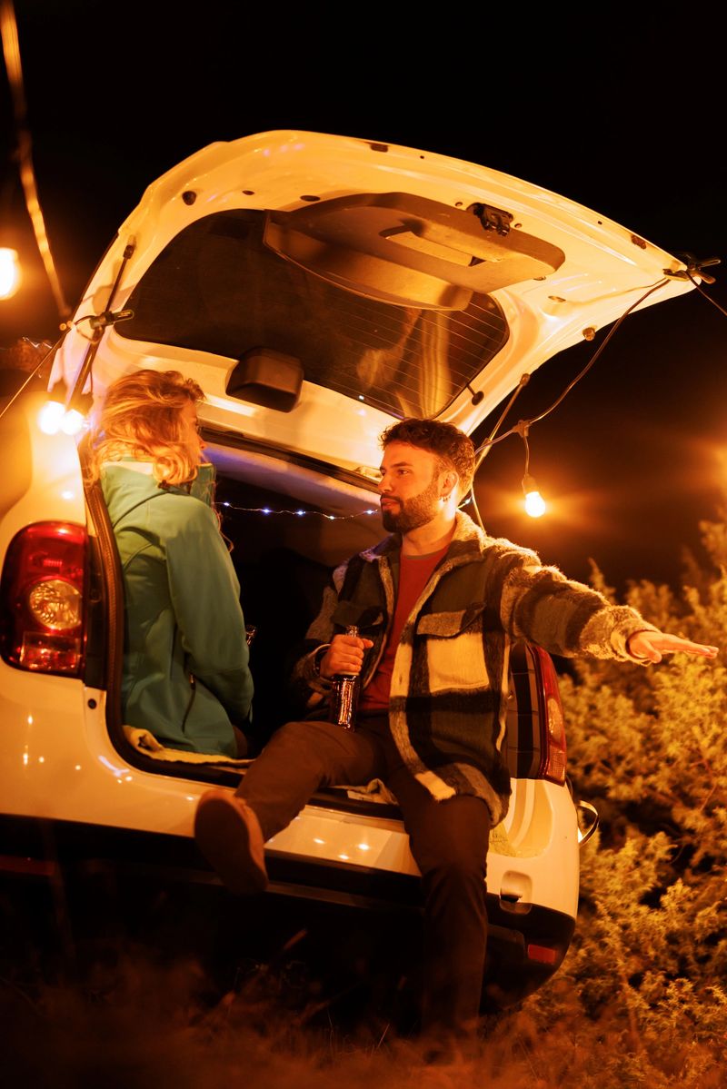 Under the glow of string lights, two friends sit at the back of an open car, sharing stories and laughter in a peaceful outdoor setting late at night.