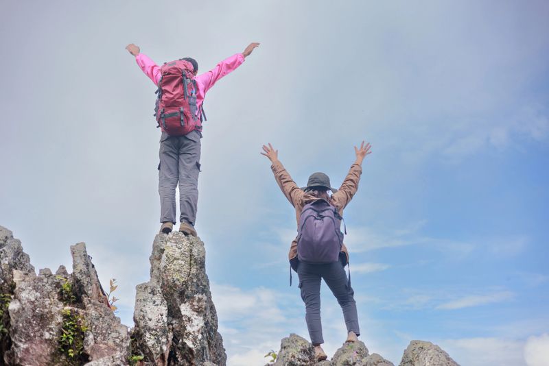 Two hikers with backpacks raising arms and trekking poles on rocky mountain peak, symbol of adventure, achievement, teamwork, freedom, success, exploration and outdoor lifestyle inspiration.