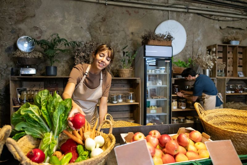 Woman organizing fresh produce while her colleague arranges dry goods in a small, local greengrocer's shop