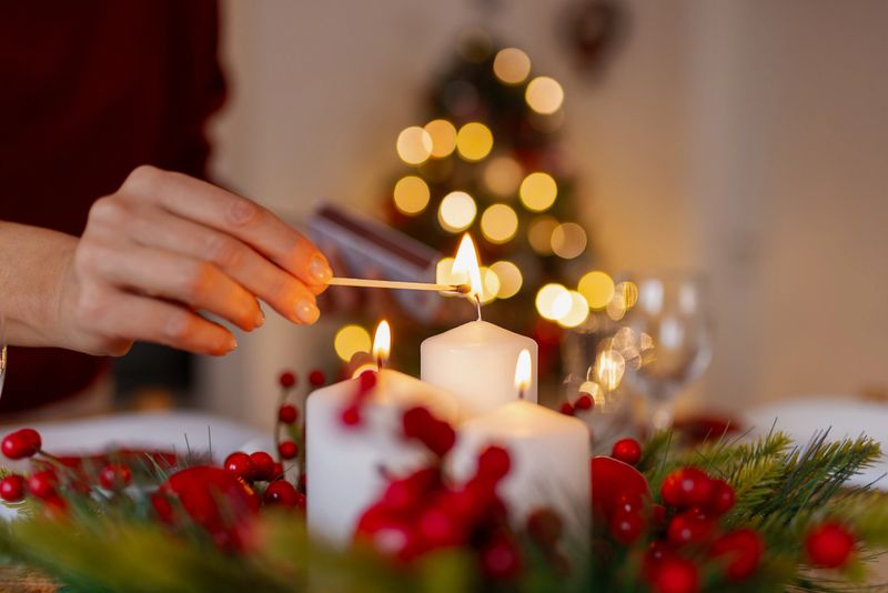 A woman's hands gently light white pillar candles nestled among red berries and evergreen sprigs on a holiday dining table. Soft bokeh lights from a Christmas tree glow in the background. Perfect for cozy holiday themes, festive decor, and romantic dinner concepts.