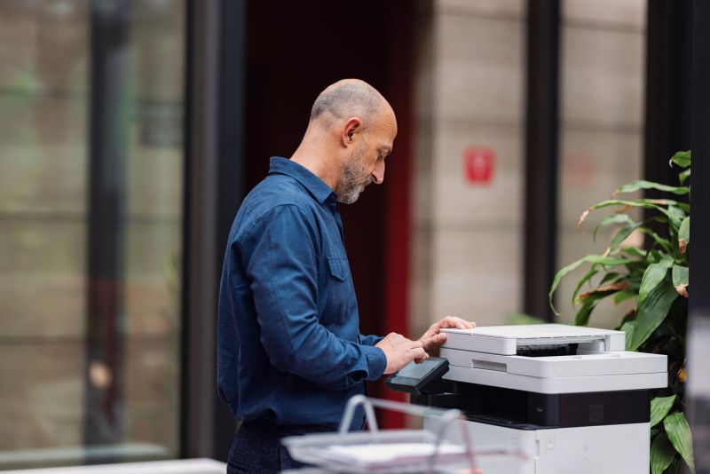 A man in a blue shirt uses a multifunction printer outside a modern office building, focusing on the touchscreen. The scene combines work, technology, and urban life with a touch of greenery.
