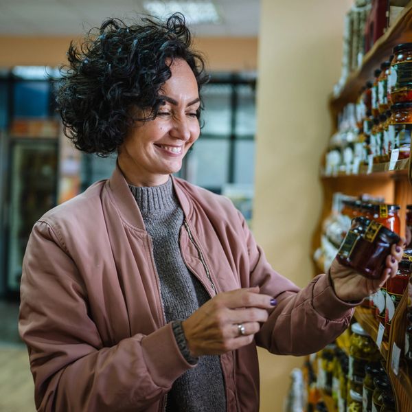 Smiling woman examines a jar on a grocery store shelf.