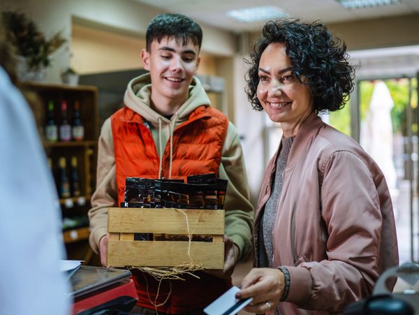 Smiling woman paying with a card while young man holds a crate of goods at a store counter.