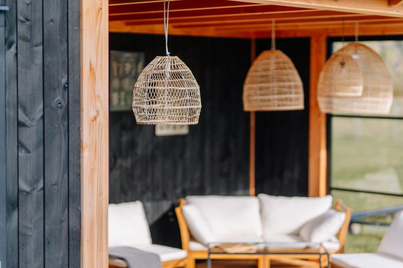 Close-up of woven rattan pendant lights hanging from a wooden ceiling in a cozy outdoor seating area with light cushions and natural decor elements.