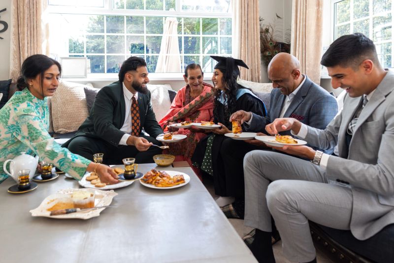 A wide shot of a multigenerational family gathered together in a well-lit living room, sharing a celebratory cup of tea and a snack to mark a graduation milestone. They are all wearing smart casual clothing. The scene captures togetherness, family support and the celebration of academic success. Located in Middlesbrough, North East England.Video of this scenario is available