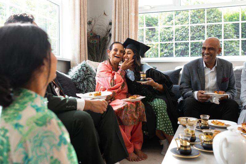 An over-the-shoulder shot of a young female adult and grandmother hugging each other in a well-lit living room, sharing a celebratory cup of tea and a snack to mark a graduation milestone, surrounded by family. They are all wearing smart casual clothing. The scene captures togetherness, family support and the celebration of academic success. Located in Middlesbrough, North East England.Video of this scenario is available