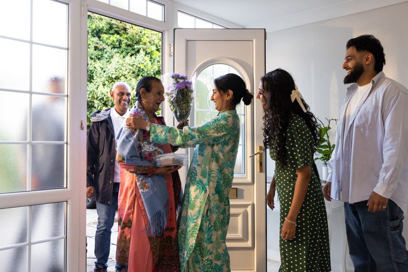 A three-quarter length family moment as relatives arrive at the front door and are greeted with smiles, hugs, and a bouquet of fresh flowers. Everyone is smiling and laughing, wearing smart casual clothing. This image beautifully captures love, connection, and the welcoming atmosphere of family gatherings.Video of this scenario is available