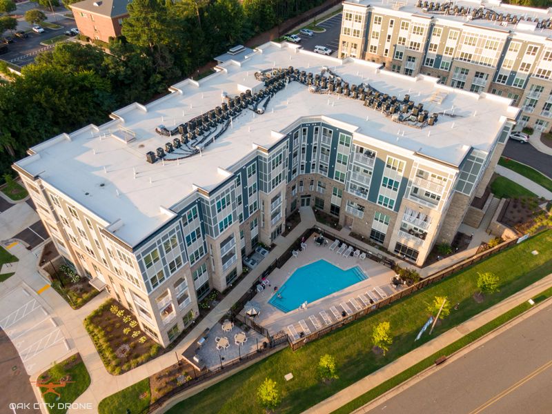 A drone aerial view of a modern multi-story apartment complex featuring contemporary architecture, a central swimming pool, landscaped grounds, and rooftop HVAC systems in Raleigh, North Carolina. The image highlights urban residential living, real estate development, and modern apartment design in a growing city.