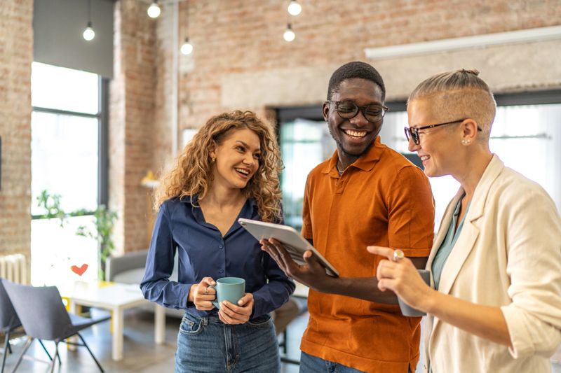 Diverse business professionals smiling and discussing work while looking at a digital tablet, fostering collaboration and teamwork in a creative, open-plan office environment