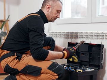 Technician in orange overalls fixing a radiator near a window.