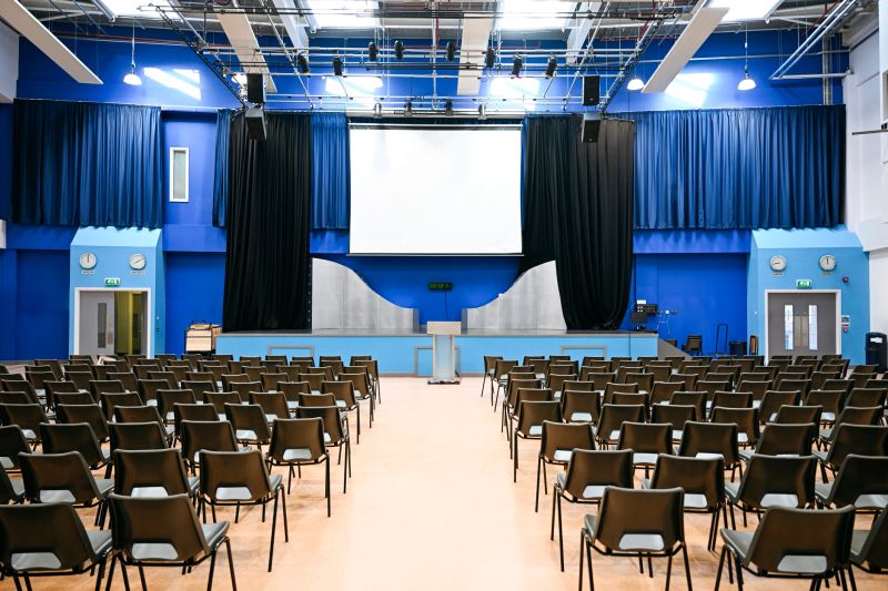 Large school assembly hall with rows of chairs, a stage and large screen without people. This is a wide shot of a large auditorium or conference room set up for a presentation or event. Rows of empty black chairs fill the foreground, leading up to a stage at the back. The stage features a white screen, flanked by blue walls draped with black curtains. A lectern stands to one side of the stage. The walls are a vivid blue, contrasting with the lighter flooring.