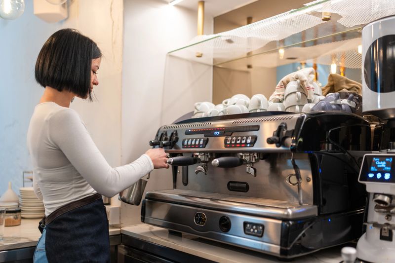 A focused young Caucasian female barista, wearing a white top and apron, foams milk at a modern espresso machine in a brightly lit cafe.