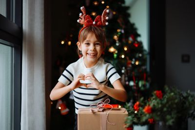 Smiling girl with reindeer antlers makes a heart with her hands by a Christmas tree and gift.