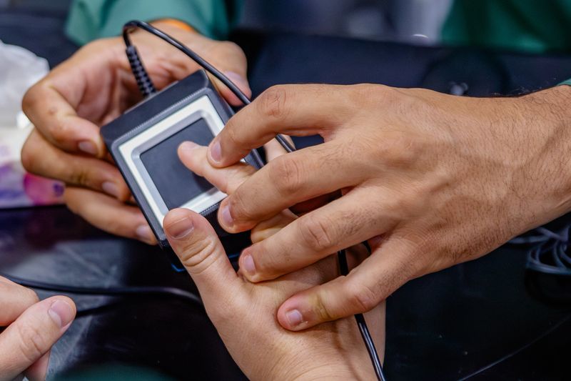 A close-up view captures two individuals engaged in a biometric scanning process. One person, wearing a green shirt, holds a square fingerprint scanner, guiding the other person's hand to place a finger on the device. The focus is on their hands and the scanner, highlighting the interaction. In the background, elements like a hand sanitizer bottle, a tissue box, and various cables suggest an office, clinic, or administrative setting. The image emphasizes security, identification, and modern technology.