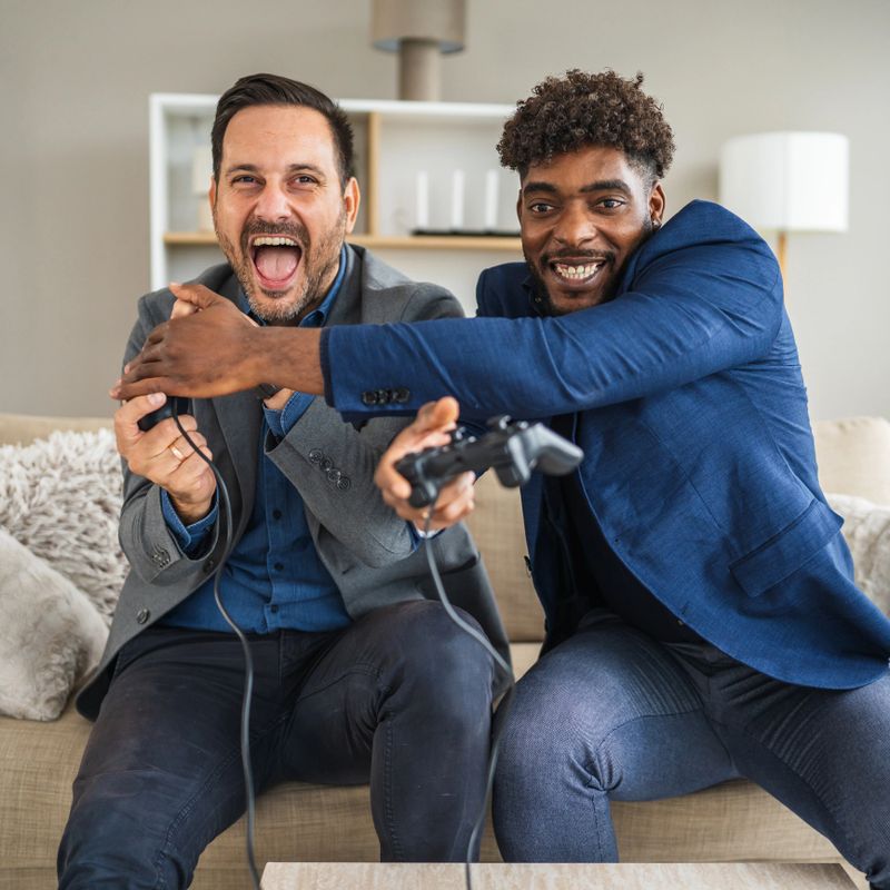 Two diverse adult men friends in business casual attire sitting on a sofa, happily competing and laughing while playing a video game console with controllers in a bright living room