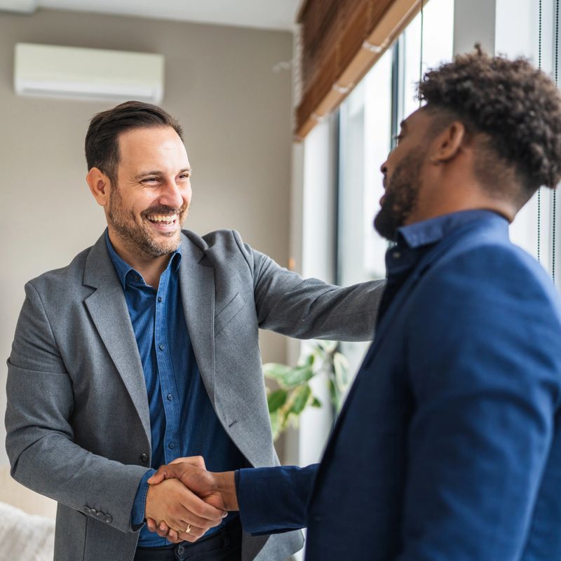 Two diverse businessmen concluding a partnership agreement, shaking hands and smiling cheerfully during a professional meeting in a modern office, symbolizing collaboration and success