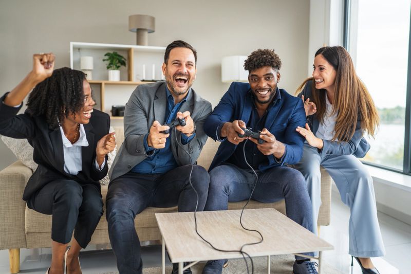 Diverse business colleagues sitting on a couch, enthusiastically playing video games during an office break, fostering team building, friendship, and collaboration