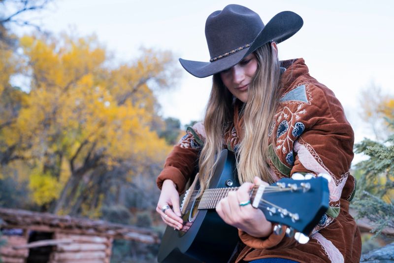 Environmental portrait of a young woman wearing a Western-style embroidered jacket and turquoise ring plays a black acoustic guitar outdoors in a rustic mountain setting. Capturing the authentic spirit of country music and rural life in autumn.