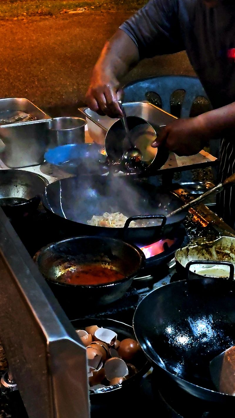 Street food vendor cooking fried noodles at night with hot woks, smoke, and bright street lights. A lively scene showing authentic Asian night market atmosphere and traditional street cooking style.