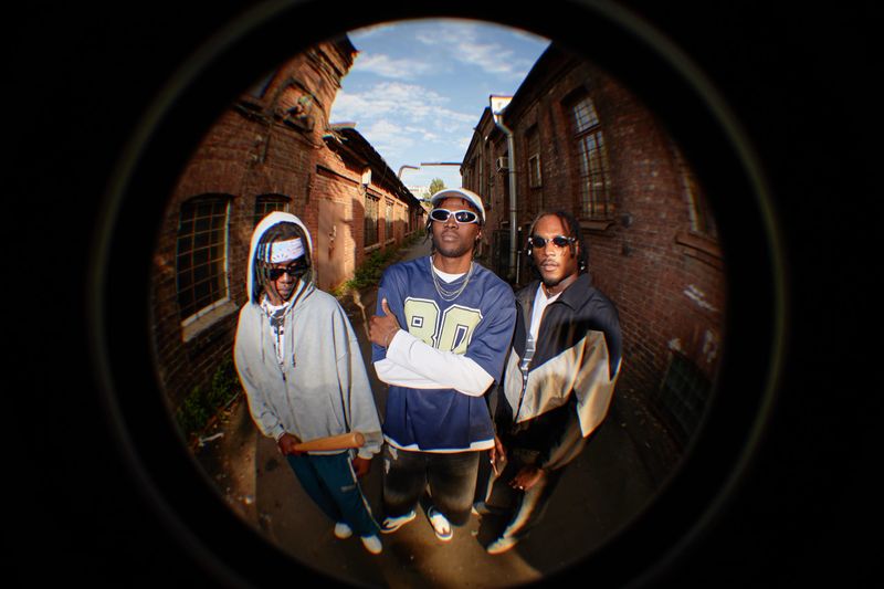 Three Black young adult men standing in urban alleyway, one crossing arms and looking confidently at camera, others wearing sunglasses and bandana, holding baseball bat