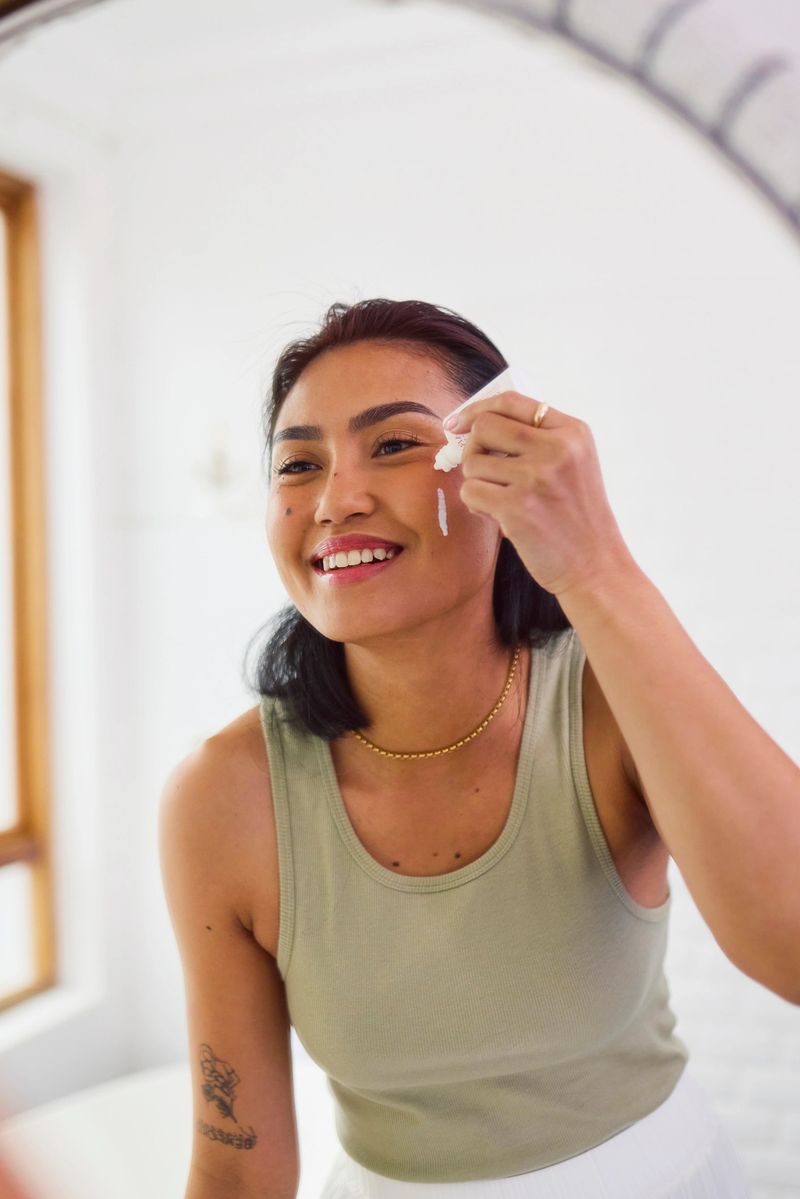 A cheerful young Asian woman smiles genuinely at her reflection, squeezing cream from a tube directly onto her cheek during her daily skincare routine in a bright bathroom.