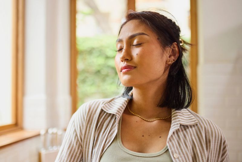 A serene portrait of a young Asian woman with her eyes closed, taking a moment for peaceful relaxation, deep breathing, or mindful meditation near a bright window indoors.