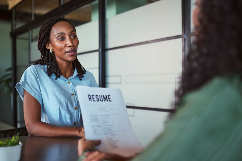 An attentive Black professional woman sits opposite an unseen interviewer, confidently speaking as the recruiter holds and reviews her paper resume across a modern office table.