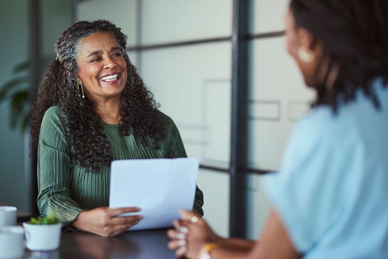 A cheerful mature Black female executive holds documents and smiles warmly while talking to an unseen candidate in a supportive and friendly professional interview or mentoring session.
