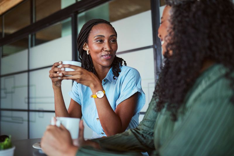 A candid shot of a young Black professional holding a coffee cup and making eye contact while intently listening to a mature colleague during an informal business discussion.