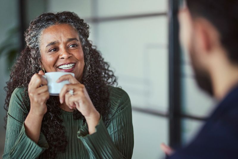 A cheerful mature Black female executive with gray curly hair holds a coffee cup and smiles warmly while actively engaging with a male colleague or mentee.