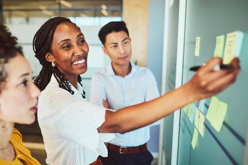 A cheerful young Black female leader writes on a glass wall covered with sticky notes, directing and engaging a diverse team during a collaborative strategy meeting.