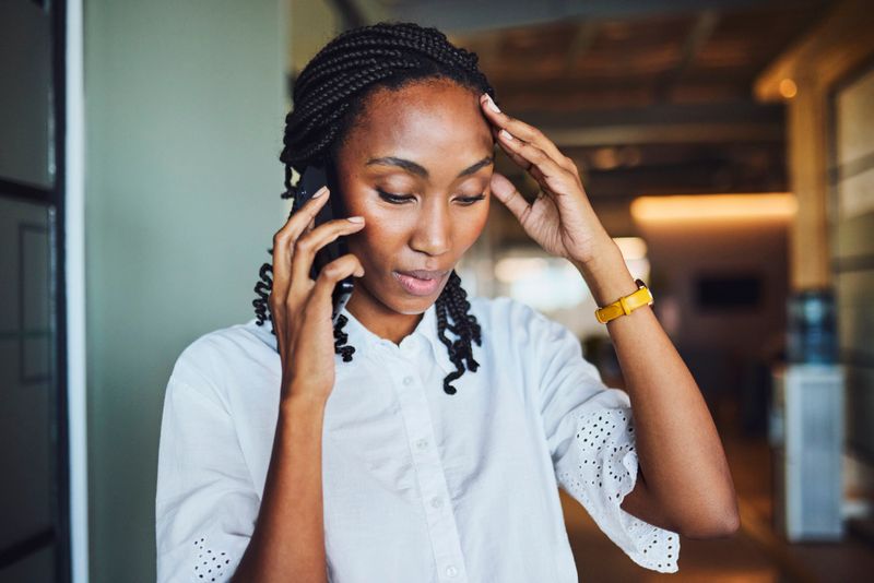 Candid close-up of a worried African American female professional on a stressful mobile phone call, holding her head in an anxious gesture in a corporate office setting.