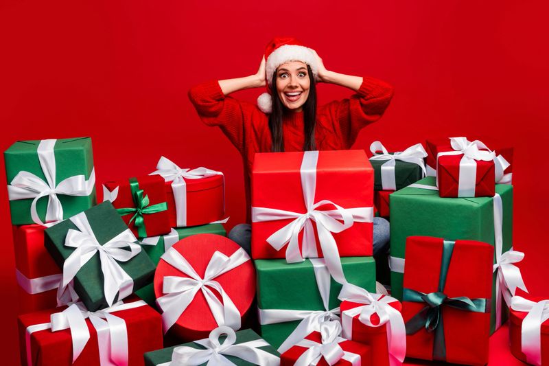 A cheerful young woman surrounded by beautifully wrapped Christmas presents, wearing a red sweater and Santa hat, expressing holiday joy.