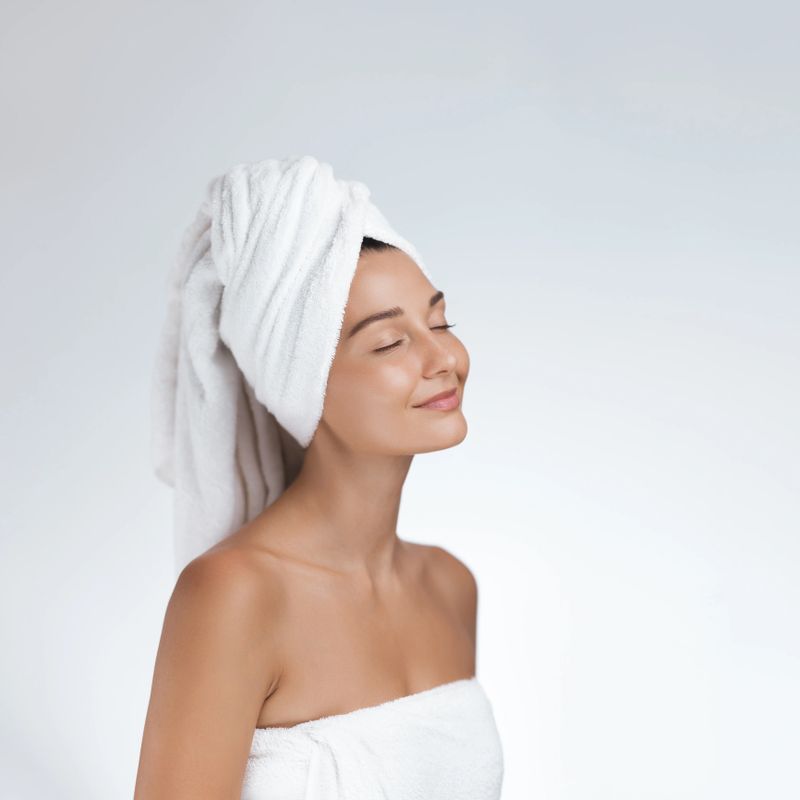 Caucasian woman wrapped in white towel with radiant skin and eyes closed, enjoying post-shower moment. High quality beauty and wellness image ideal for skincare, spa, or cosmetic promotions.