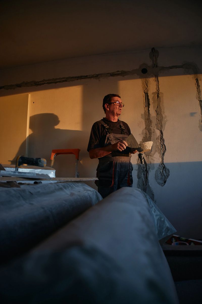 Middle-aged male contractor wearing work clothes inspects wall repairs during sunset light. Tools and construction materials are visible in an indoor setting, emphasizing renovation and craftsmanship.