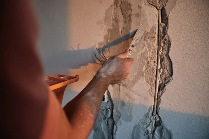 Close-up of a male worker repairing damaged plaster on a wall with a trowel. The image shows detailed texture of cracked wall surface and construction work in progress indoors with warm lighting.