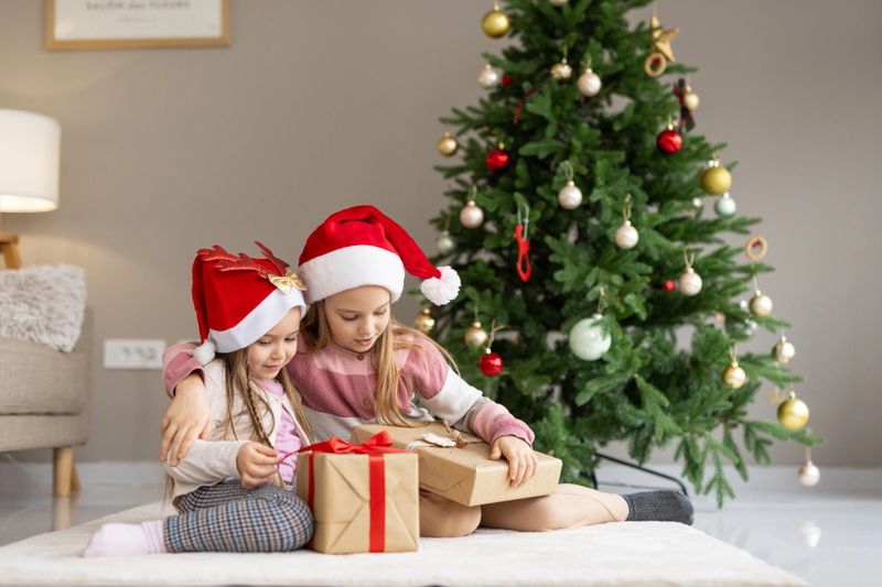 Two cheerful girls, dressed in festive attire, sit near beautifully wrapped gifts, embodying the joy and spirit of the holiday season.