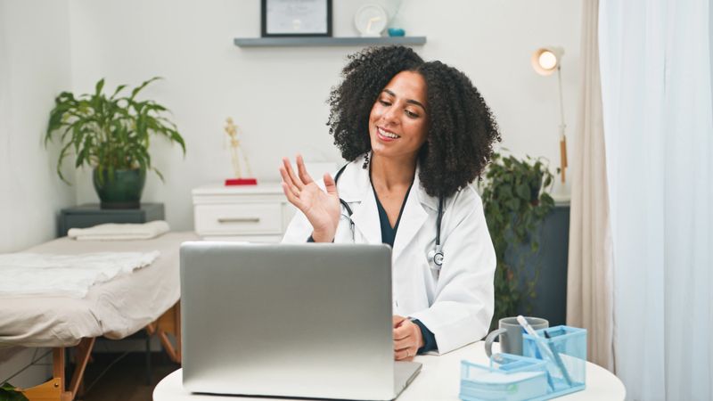 A smiling female doctor in a white coat conducts a telemedicine session with a patient via laptop in a bright, well-equipped clinic space featuring plants and comfortable furnishings.