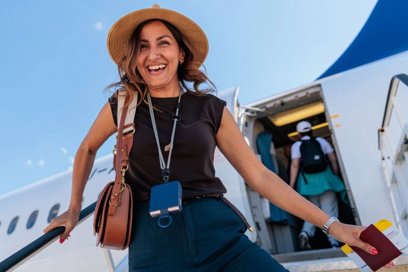 Cheerful woman wearing a hat and casual outfit smiling while walking up the stairs to board an airplane, holding her passport and plane ticket on a sunny day.